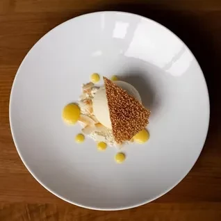 Plated dessert: creamy quenelle, sesame brittle, yellow sauce, white crumbles.