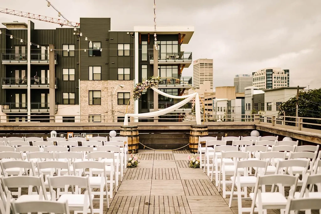 Rooftop wedding setup: white chairs, floral arch, city skyline background.