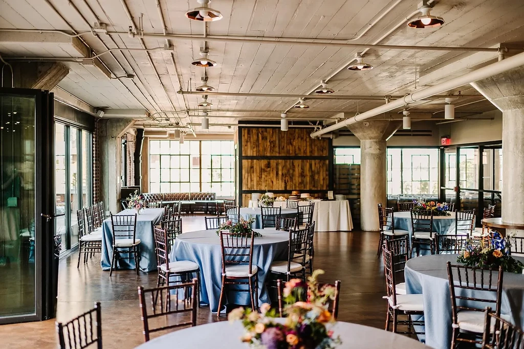 Elegant industrial-chic event hall with blue tables and floral centerpieces.
