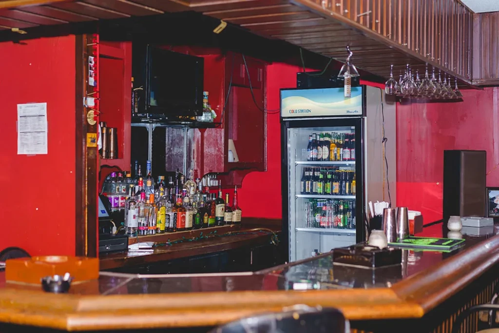 Dimly lit bar with a wooden counter, red walls, and bottled drinks.