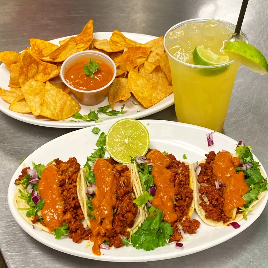 Three meat tacos, tortilla chips, salsa, and a margarita on a counter.