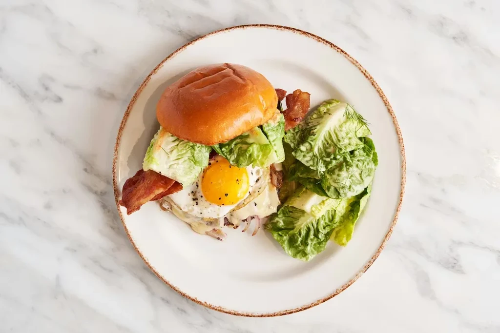 Plated brioche sandwich with crispy bacon, fried egg, cheese, and lettuce.