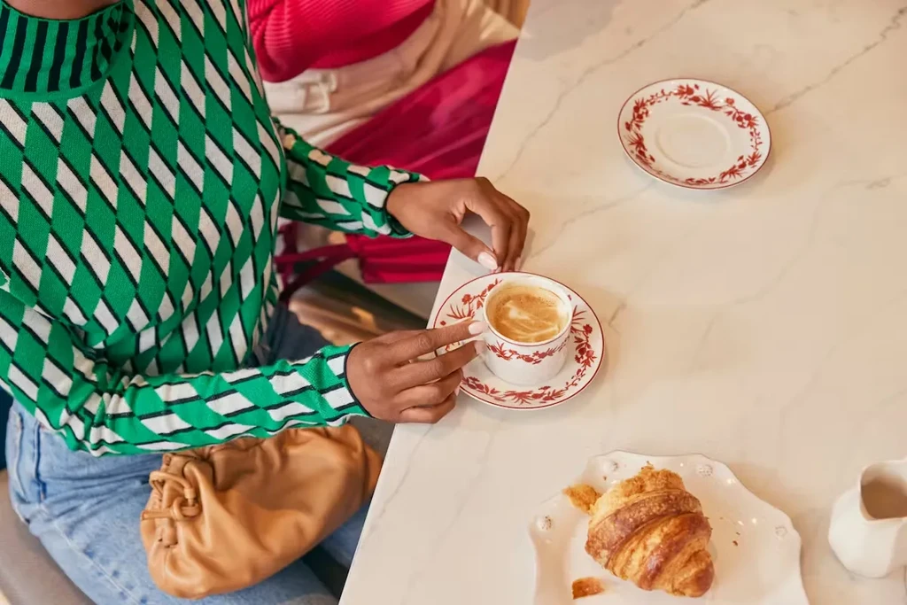 Person holds latte; croissant and floral teacups on marble table.