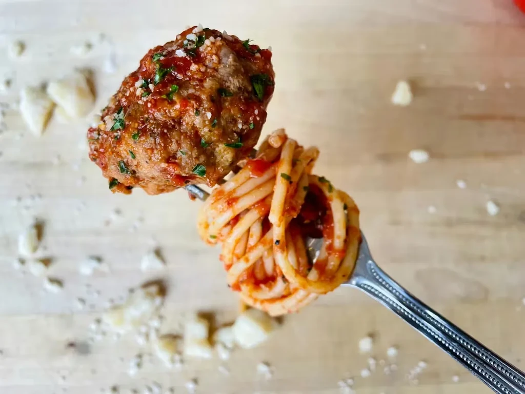 Fork lifting a meatball and spaghetti with sauce, herbs, and Parmesan.