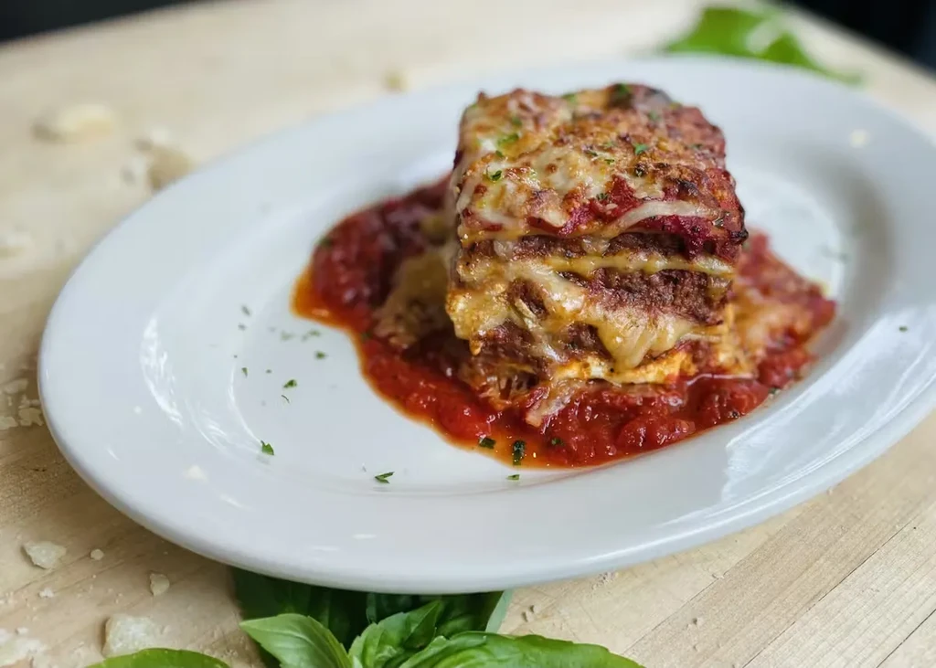 Lasagna on a white plate with layers, cheese, red sauce, and basil.