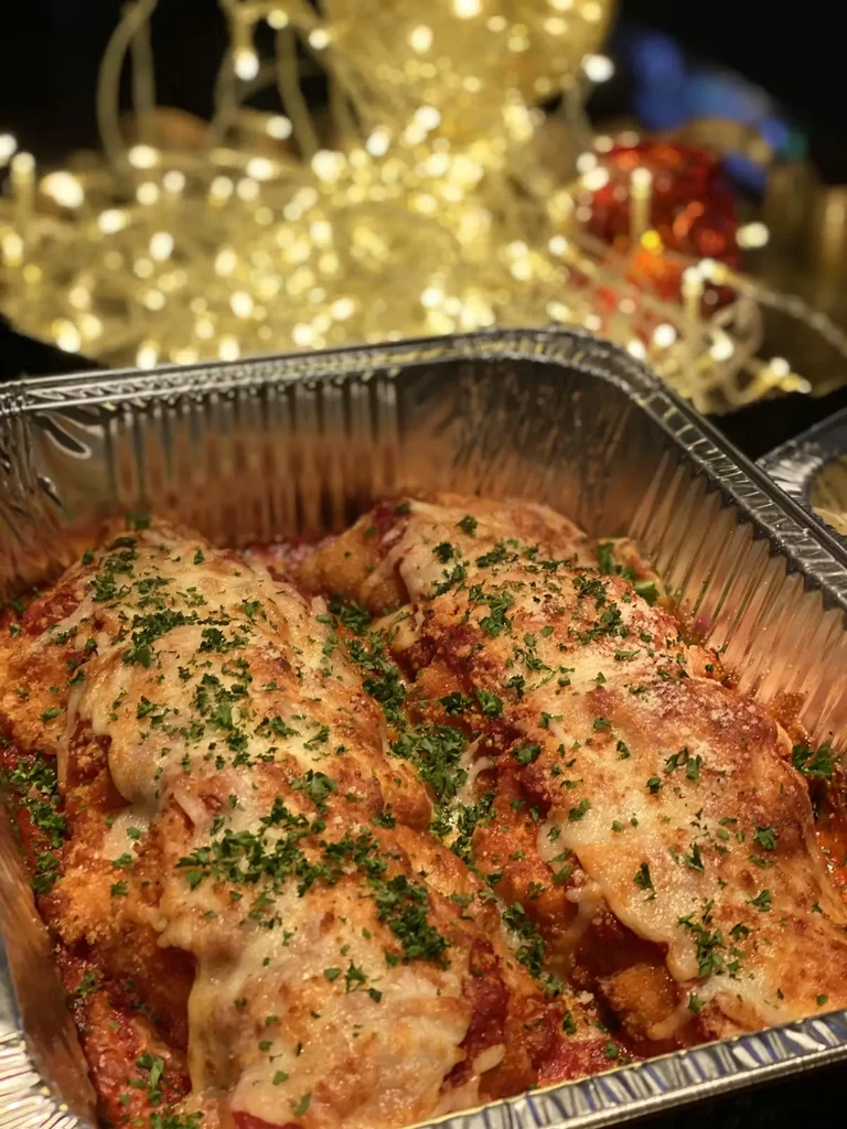 Foil container with cheesy baked dish, red sauce, herbs, festive lights.