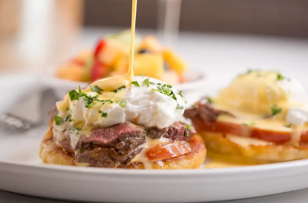 Steak Eggs Benedict with poached egg, hollandaise, herbs, and fruit.