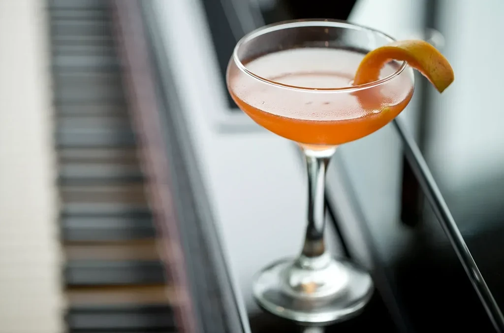 Cocktail in coupe glass with orange peel garnish, on a piano.