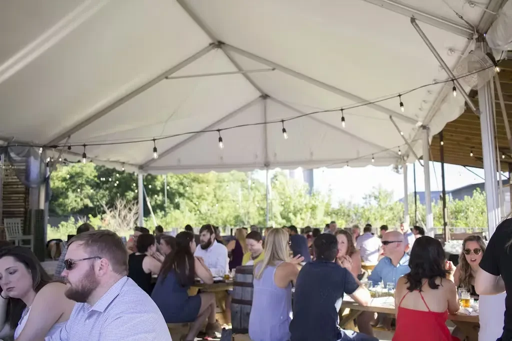 People socialize at wooden picnic tables under a large white tent.