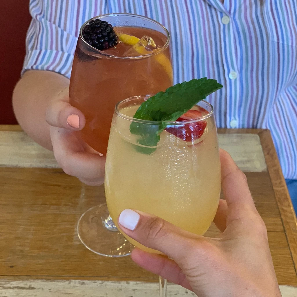 Two hands toast with cold garnished drinks, one yellow, one reddish-brown.