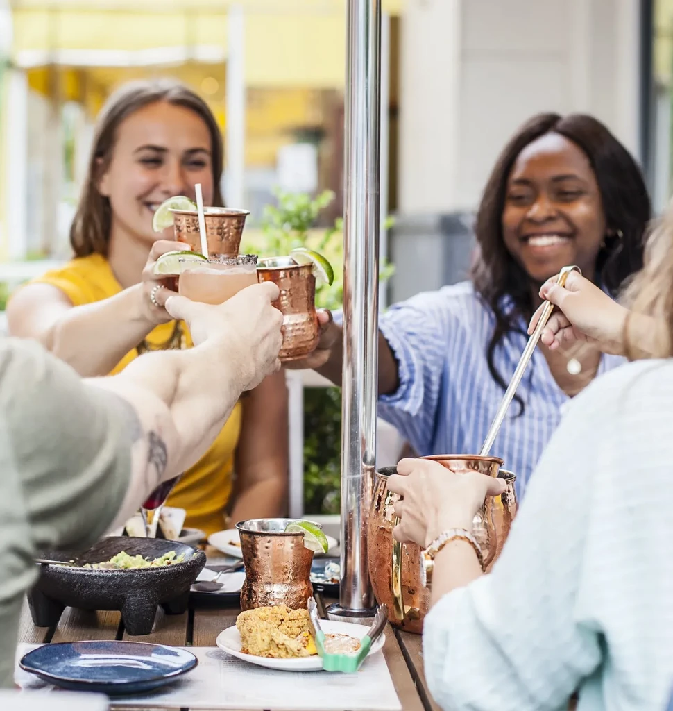 Four people clinking copper mugs and pitcher, sharing food outdoors.