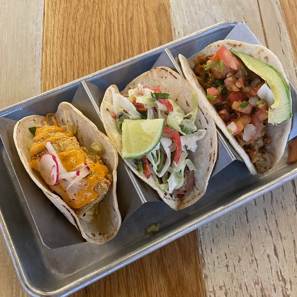 Three diverse tacos in a metal tray on a light wooden table.