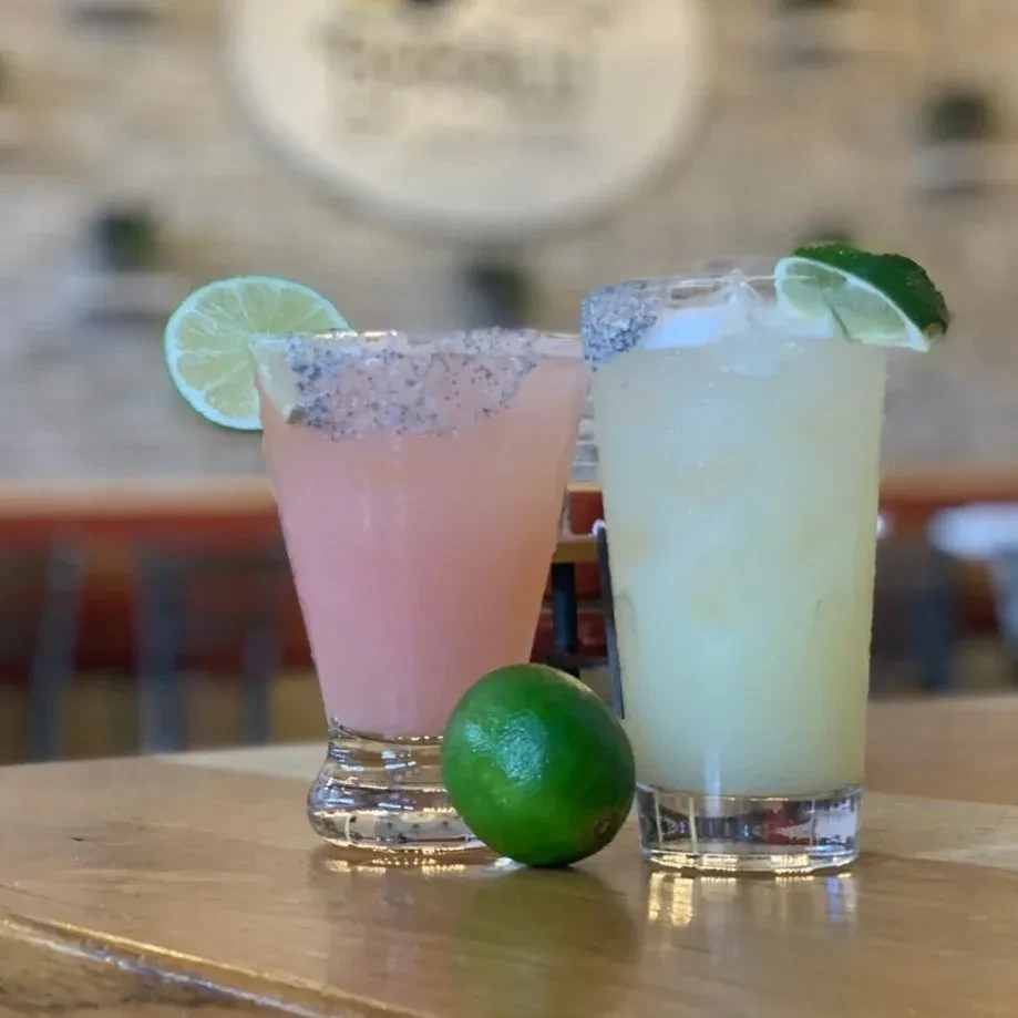 Pink and green cocktails with dark rims and lime on a table.