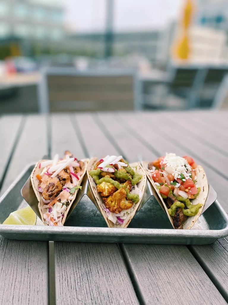 Three diverse tacos in a metal holder, lime wedge, outdoor setting.