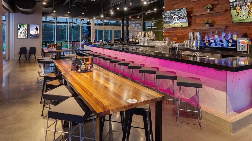 Spacious modern restaurant bar, communal table, pink-lit counter, sports TVs.