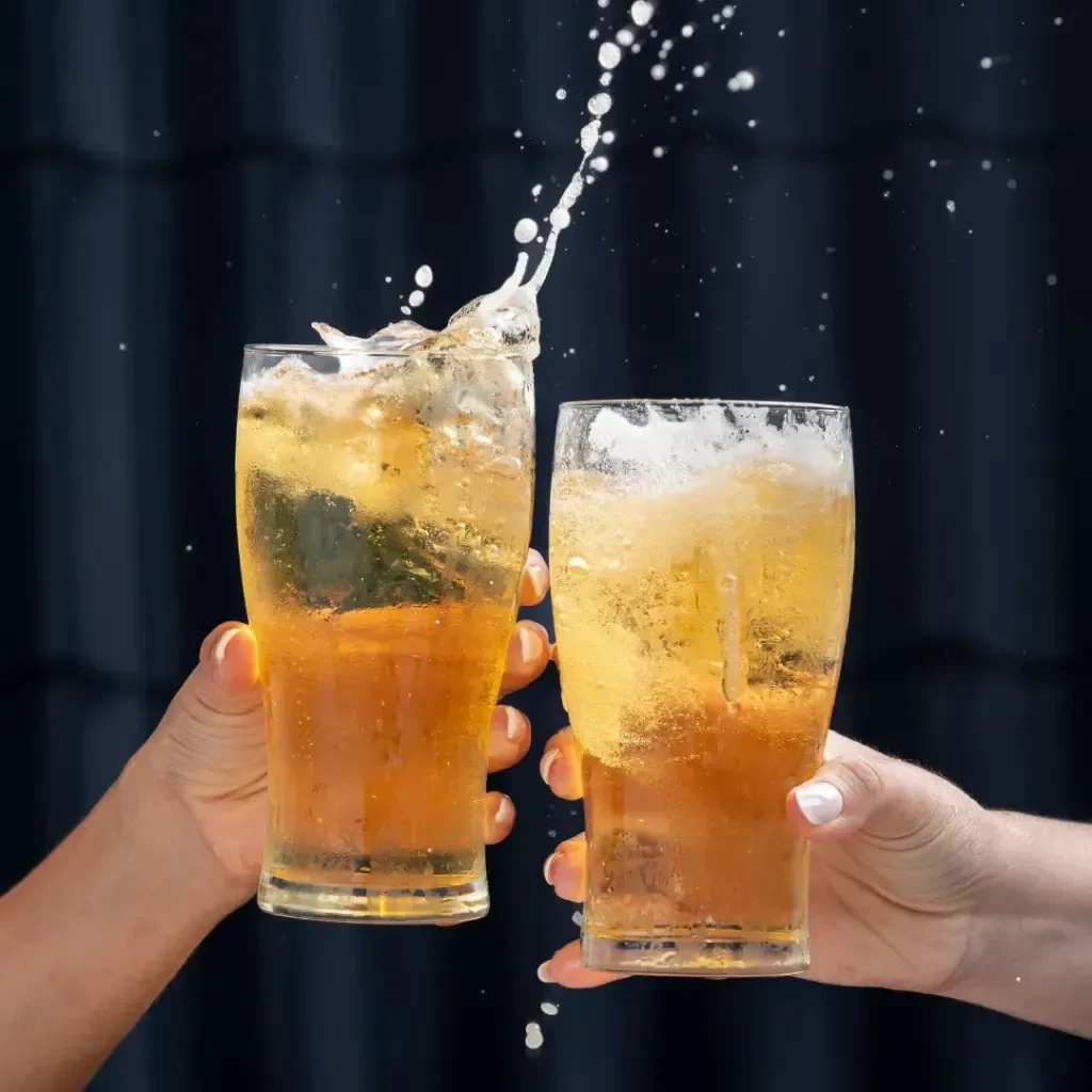 Two hands clink glasses of golden sparkling beverages, splashing.