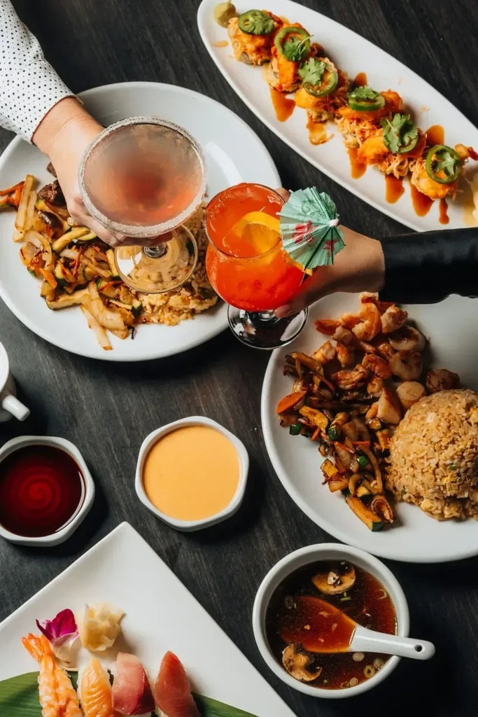 Hands hold cocktails above a vibrant table of Japanese sushi and hibachi.