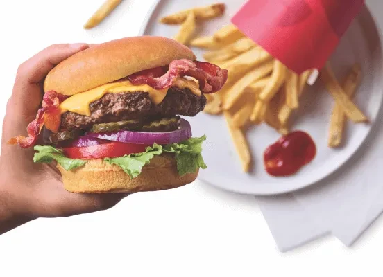 Hand holds a cheeseburger with bacon, melted cheese, and vegetables. Fries nearby.