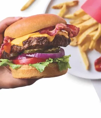 A hand holds a tall beef burger with cheese, bacon, and fries.