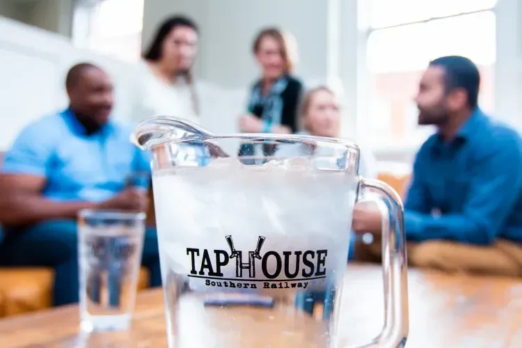 Ice water pitcher with Tap House logo, blurred people socializing.