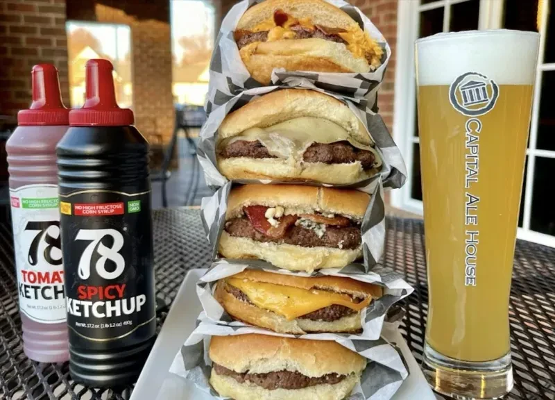 Stack of five wrapped cheeseburgers, ketchup, and beer on an outdoor table.