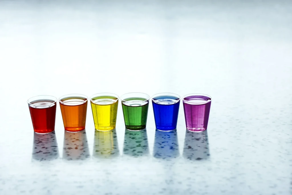 Six shot glasses with rainbow liquids on a reflective surface.