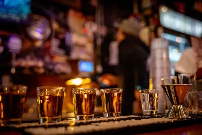 Three amber shots, jigger, and a blurred bartender at a bar.