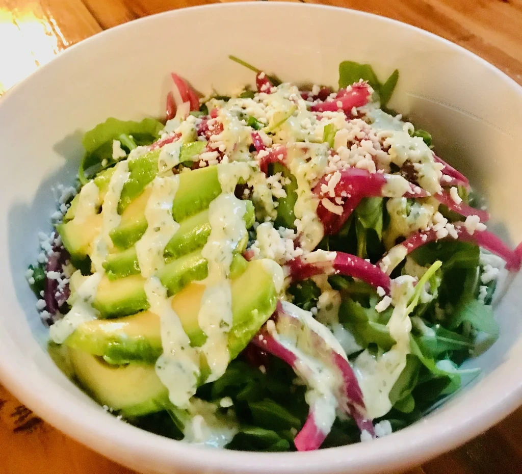 Salad in white bowl: greens, avocado, pickled onion, crumbly cheese.