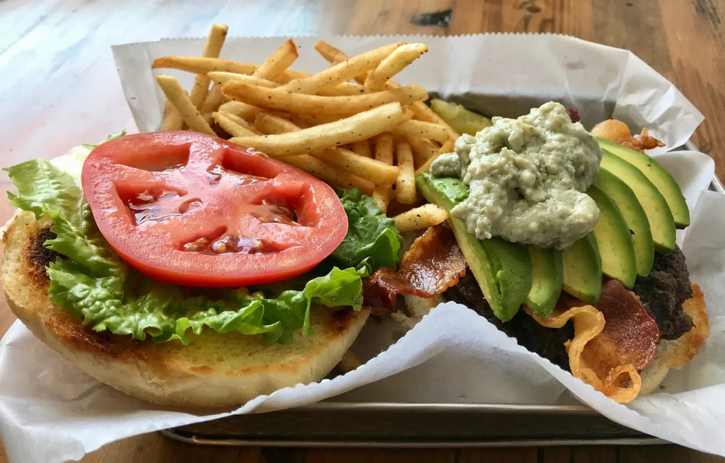 Open-faced burger with bacon, avocado, and seasoned fries on a tray.