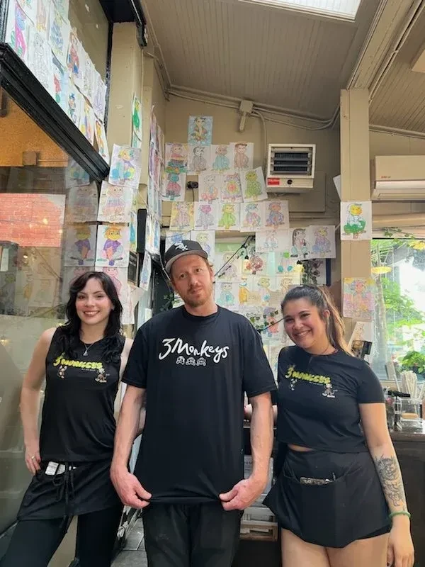 Three individuals smile, wearing '3 Monkeys' shirts, against vibrant children's art.