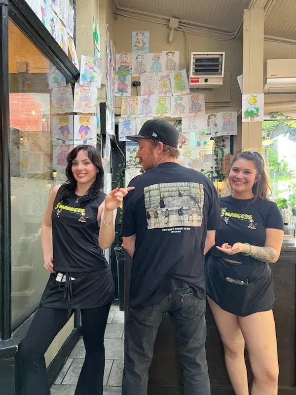 Three restaurant staff stand before a wall of colorful children's art.
