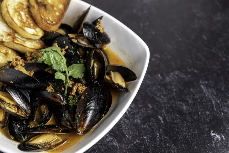 Steamed mussels in white bowl with amber broth, chorizo, cilantro, bread.