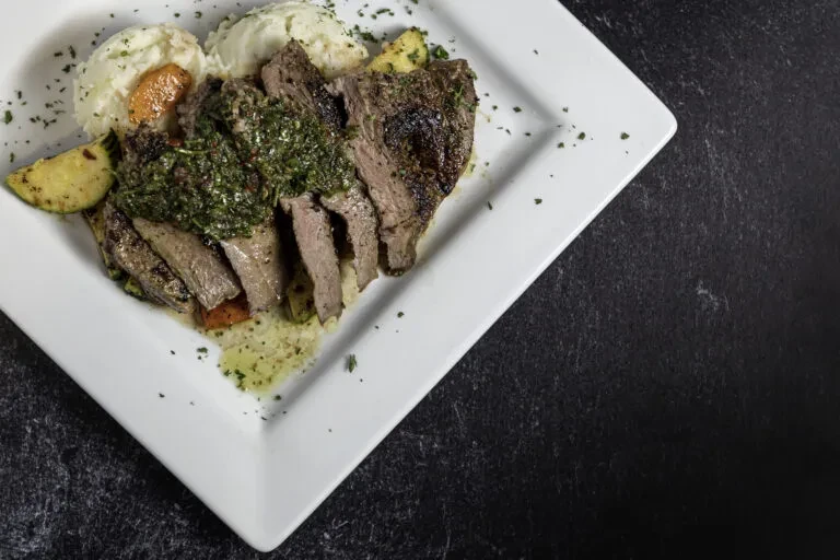 Sliced steak with chimichurri, mashed potatoes, and grilled vegetables.