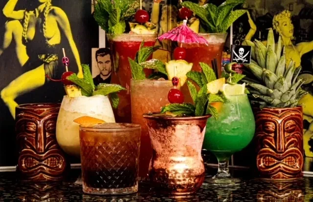 Seven colorful, tropical cocktails on a bar with retro themed art.