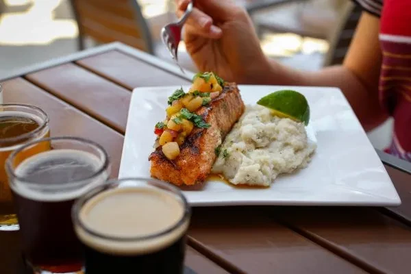 Hand with fork over grilled fish, fruit salsa, mashed potatoes, beers.