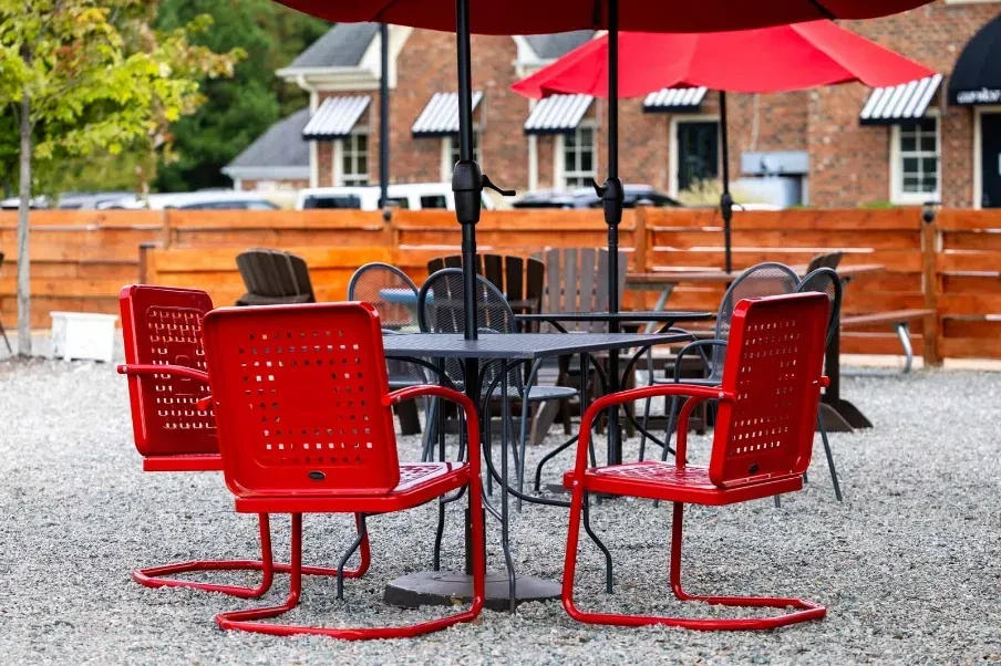 Outdoor gravel patio with red metal chairs, dark tables, red umbrellas.