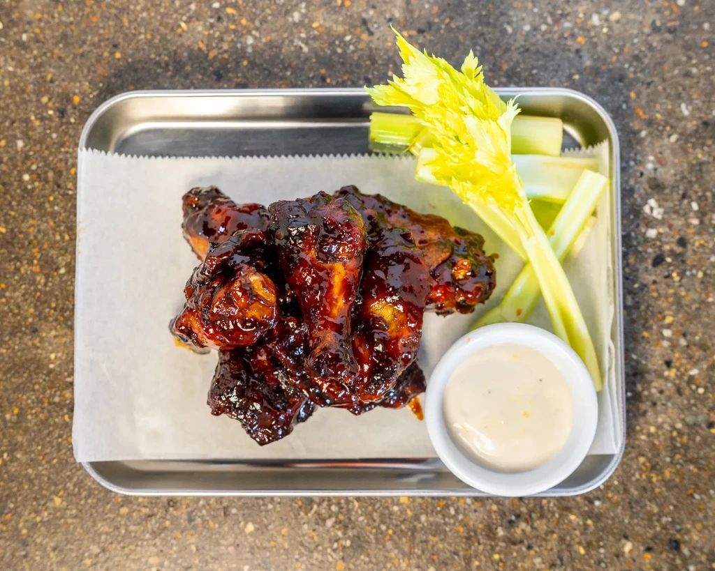Deeply glazed chicken wings, celery, and dipping sauce on a metal tray.