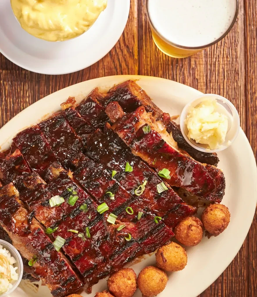 Barbecued ribs, hushpuppies, coleslaw, and beer on a rustic wooden table.