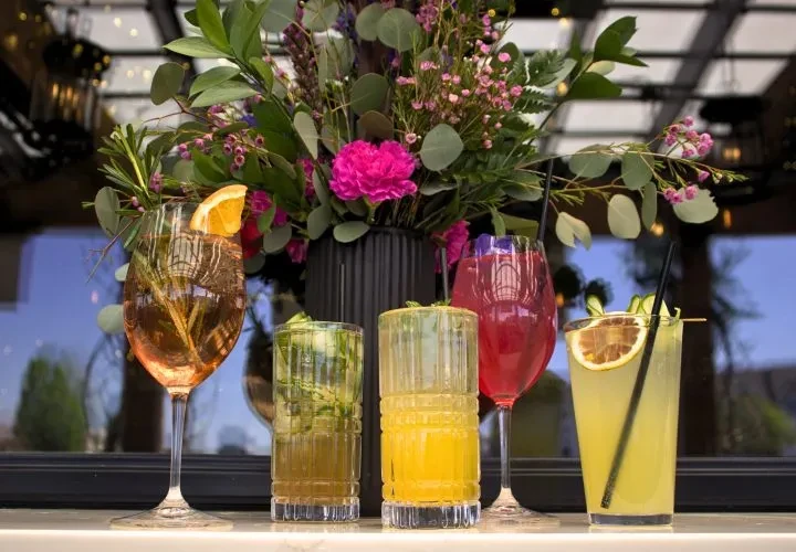 Five colorful, garnished drinks on a counter with flowers.