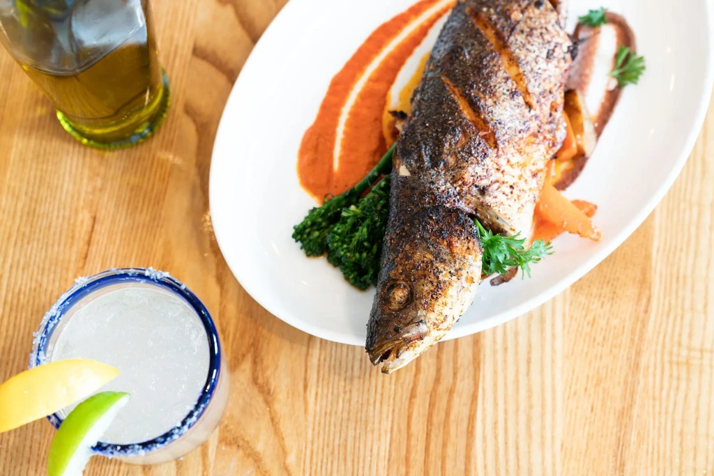 Whole grilled fish, sautéed vegetables, sauce, and a cocktail on a table.