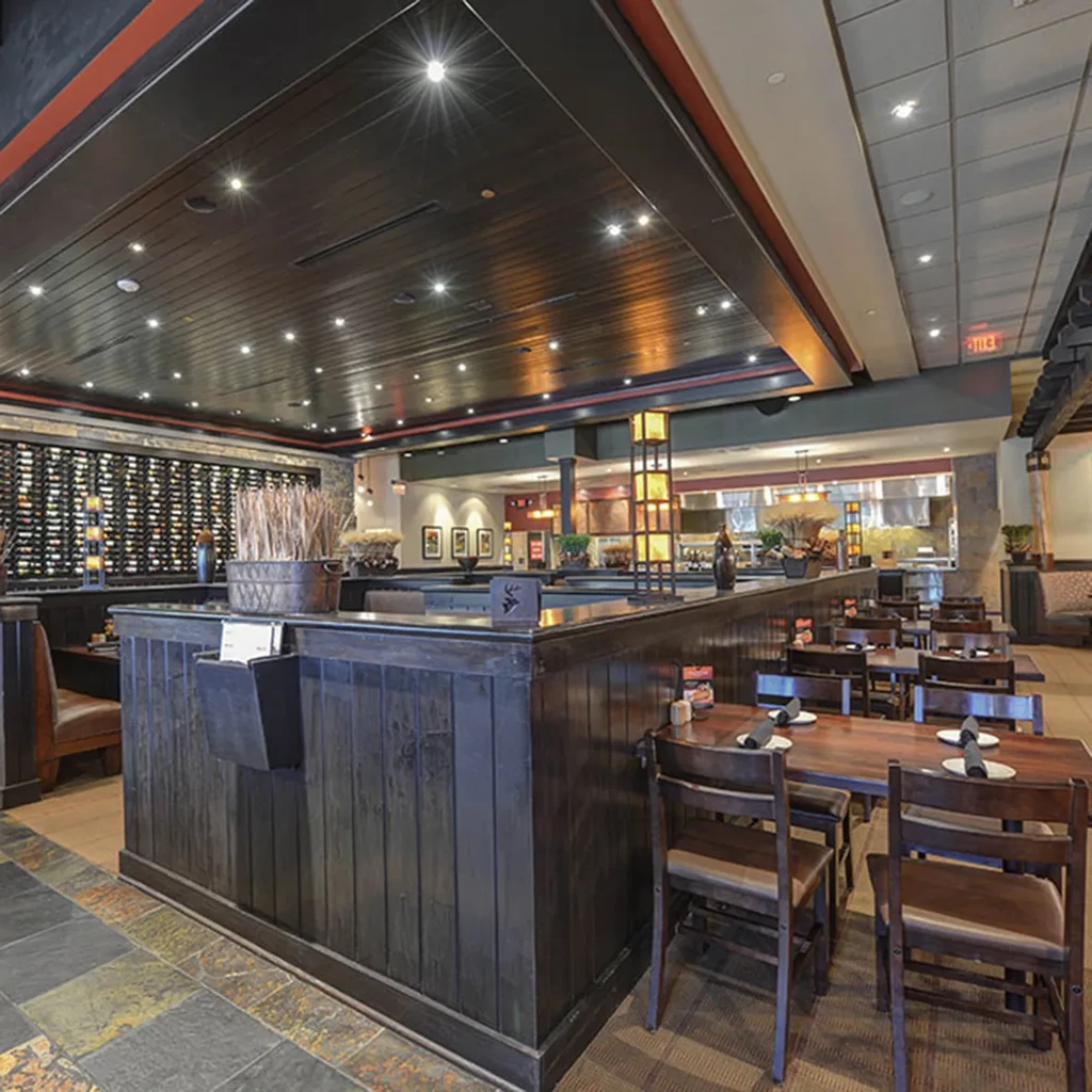 Restaurant interior: dark wood, set tables, wine rack, open kitchen, warm lighting.