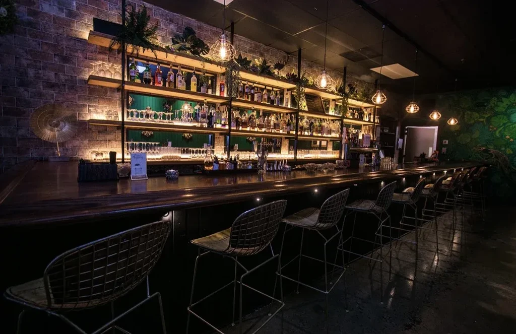 Restaurant bar with dark wood counter, lit liquor shelves, and leafy mural.