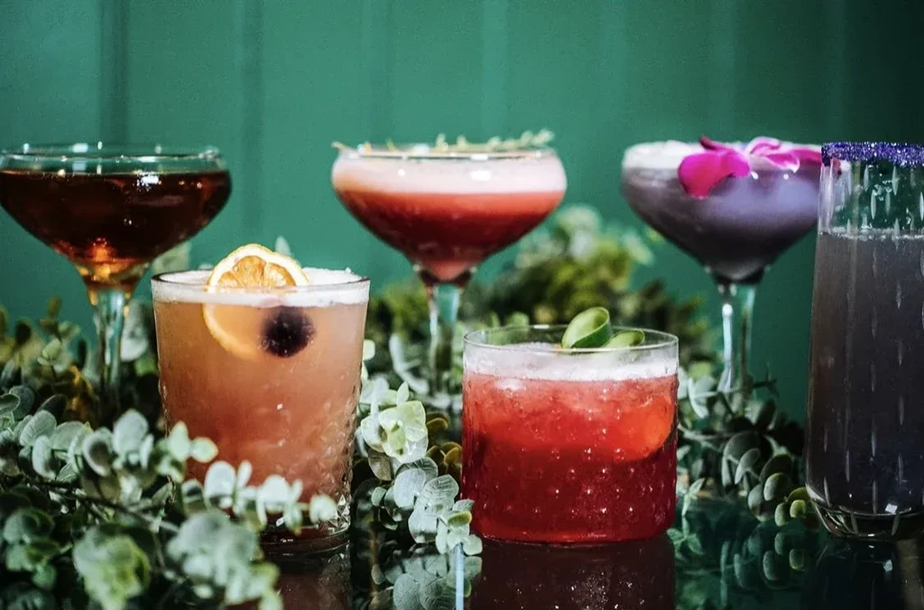 Five distinct, colorful cocktails with various garnishes and green foliage.