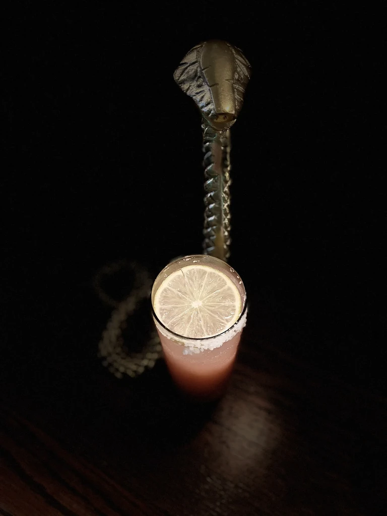 Spotlit reddish-pink cocktail with lime, salt, and a metallic cobra.