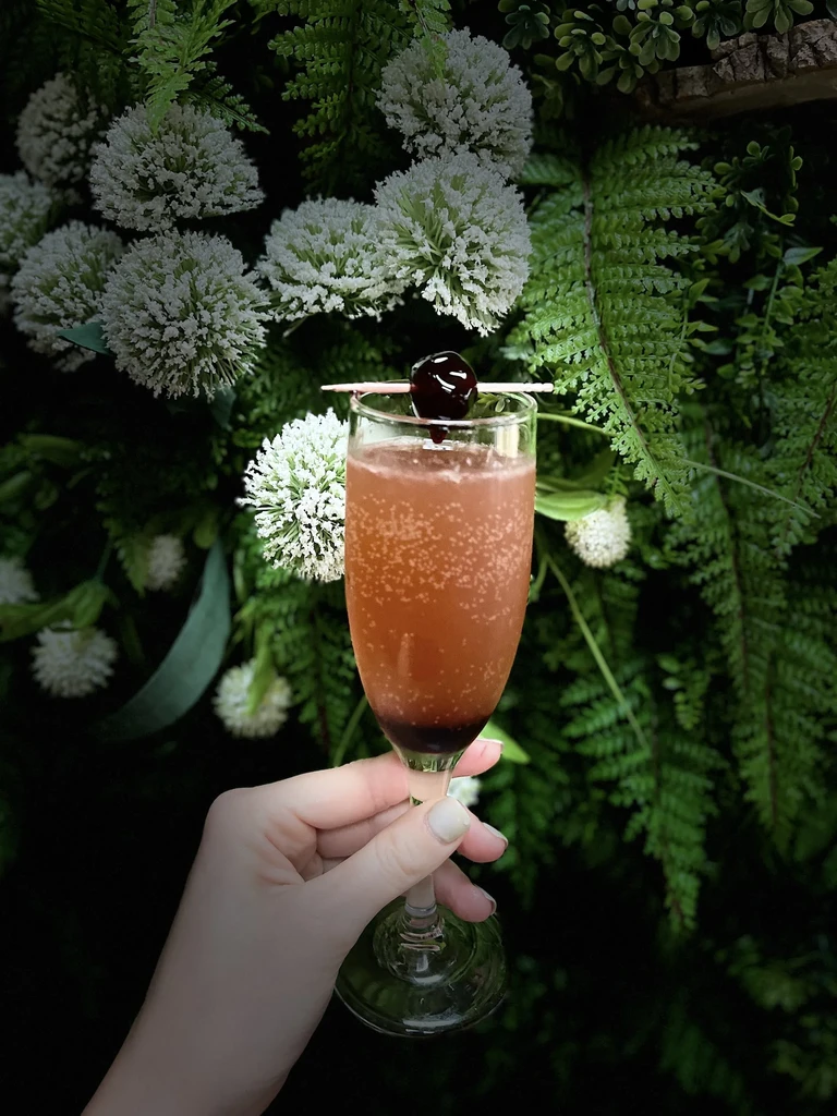 Hand holding a reddish-brown cocktail with a cherry against blurred green foliage.