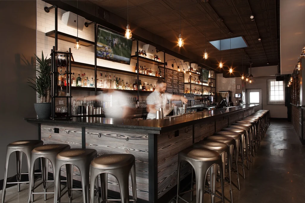 Modern industrial-chic bar interior with long counter, stools, and active bartender.