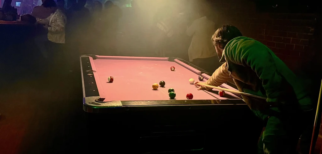 Person in green jacket playing pool on a vibrant pink table.