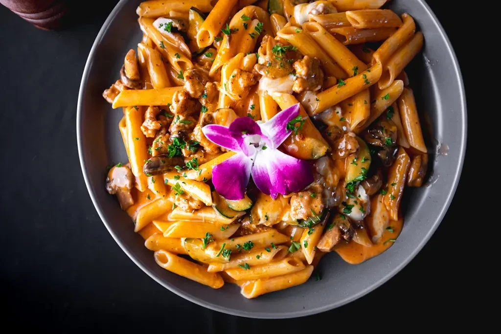 Creamy penne pasta with vegetables, protein, and purple orchid garnish.