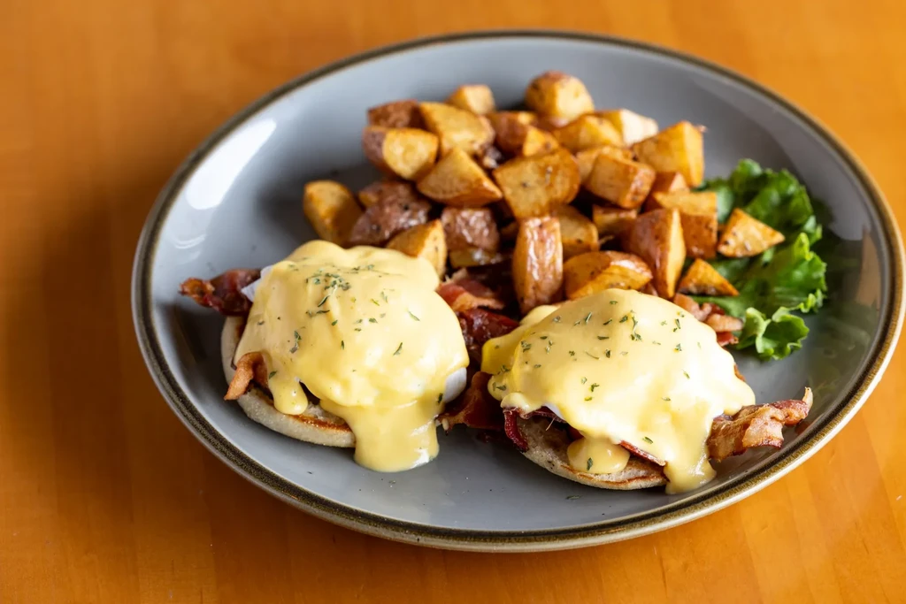 Two Eggs Benedict with bacon, hollandaise, and home fries on a plate.
