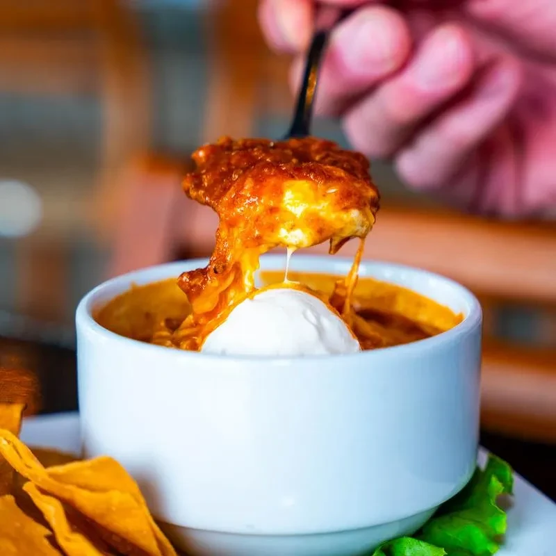 Close-up: Spoon lifts gooey cheese from chili dip with tortilla chips.